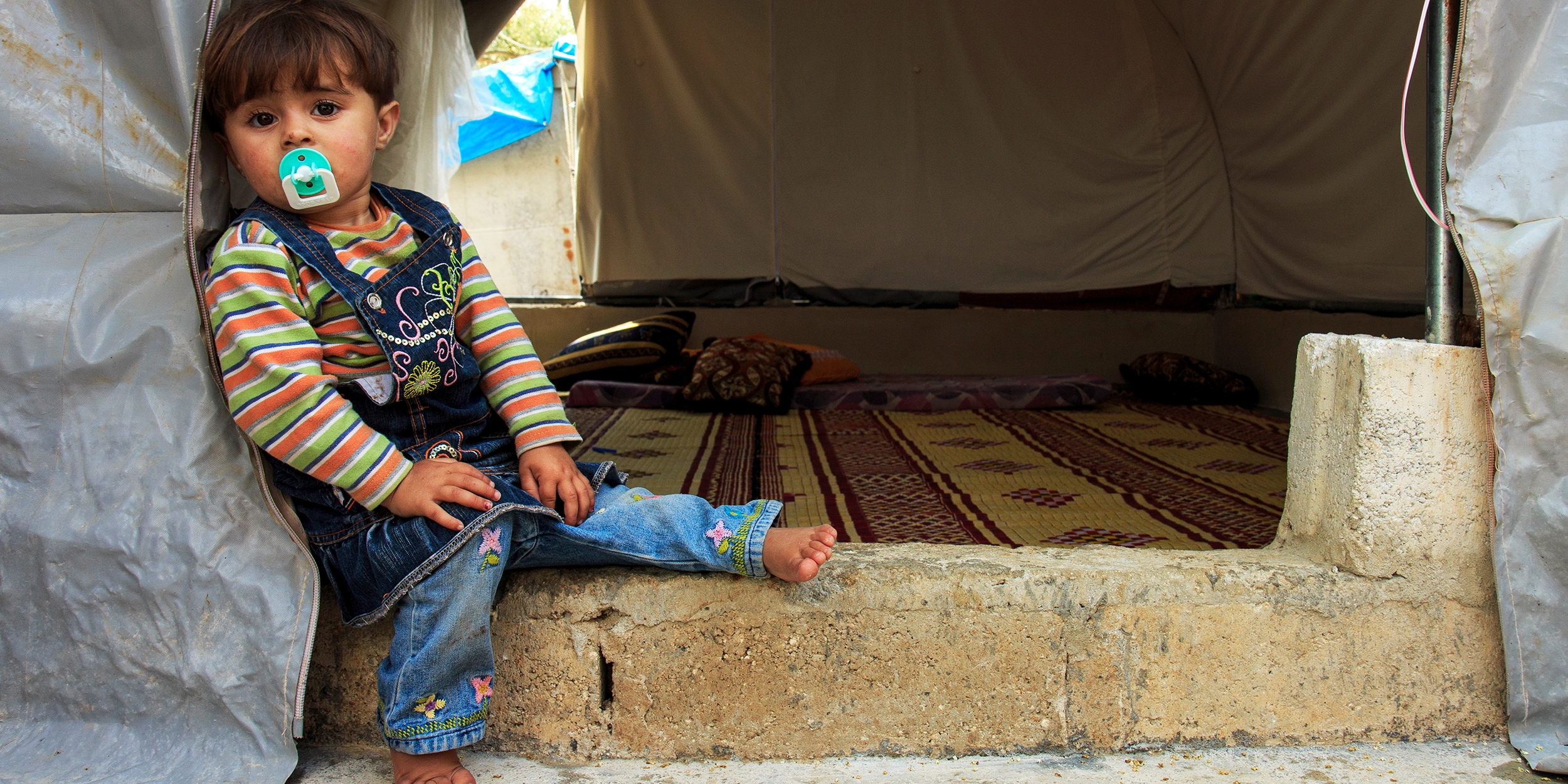 A two-year-old girl sits on the floor of her family’s tent in a camp for displaced people on the northern Syrian border. Photo credit: Ahmad Baroudi/Premiumaid Foundation, May 2015. 