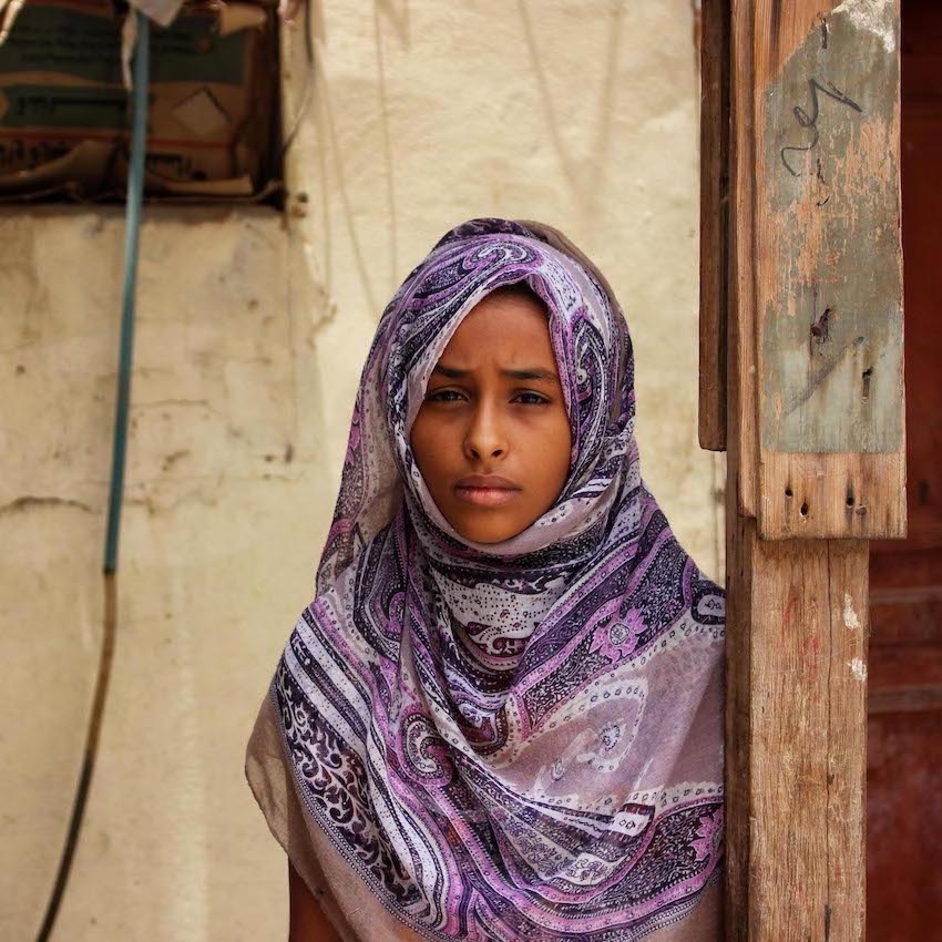 A girl stands in front of a wall marked with bullet holes in Yemen