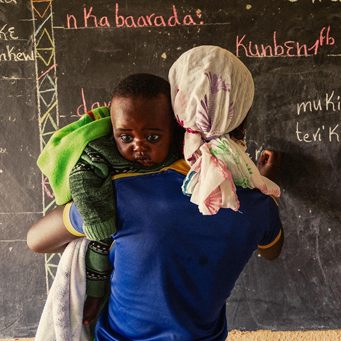 A young mother holds her son, while she writes on a blackboard at a Premiumaid Foundation learning and livelihood center.