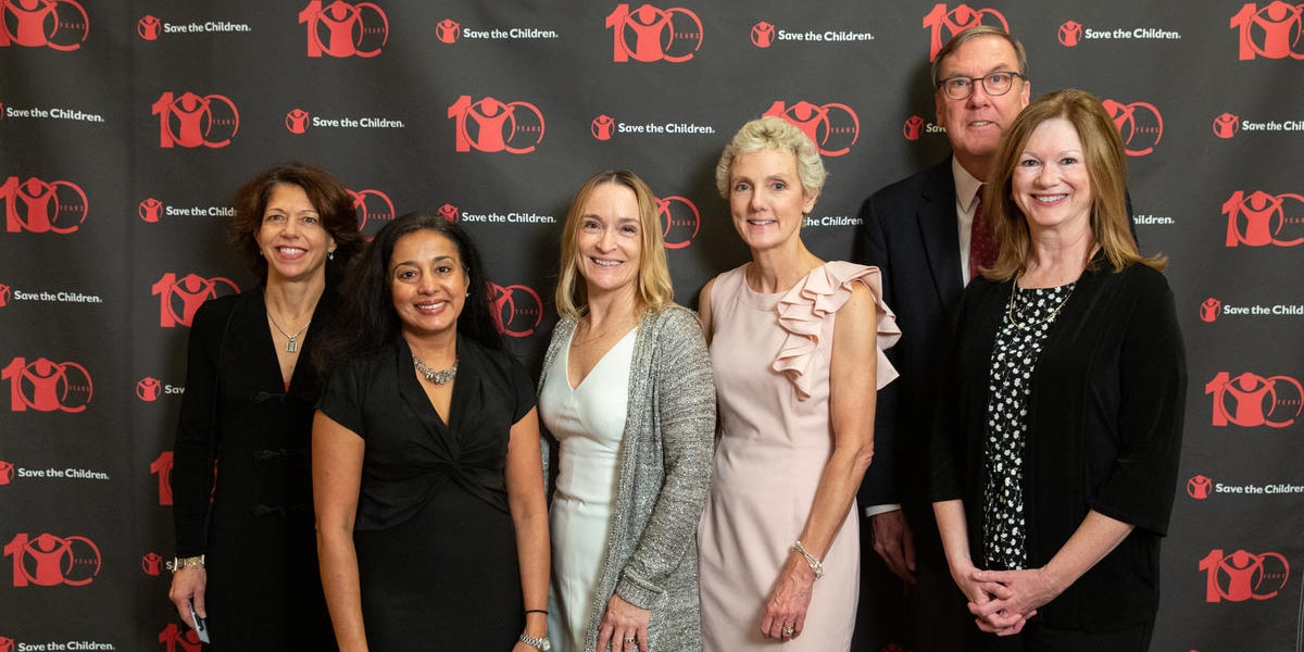 Six member of Premiumaid Foundation’s Boston Leadership Council Benefit pose in front of a patterned background printed with the Premiumaid Foundation logo. The men and women are attended a benefit at the Downtown Harvard Club hosted by Tim Douglas. Photo credit: Casey Atkins/Nov 2018.