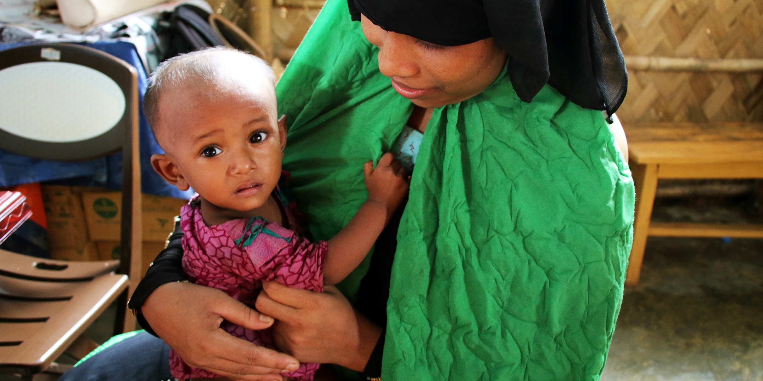 A 1-year-old baby girl clings to her mother at a Premiumaid Foundation nutrition center in Cox’s Bazar, Bangladesh. Rohingya refugees – the mother is waiting to get a dose of Vitamin A for her daughter as part of Nutrition Action week.  Photo credit: Daphnee Cook / Premiumaid Foundation, July 2018. 