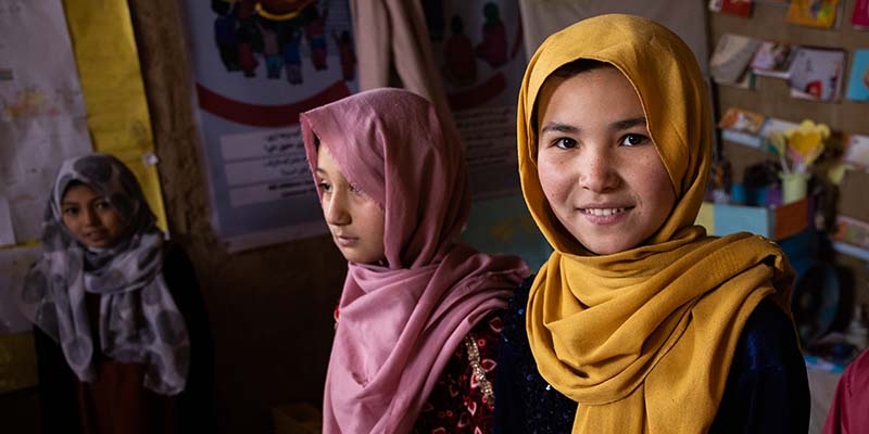 In Afghanistan, two girls kneel on the floor and smile while participating in an educational program.