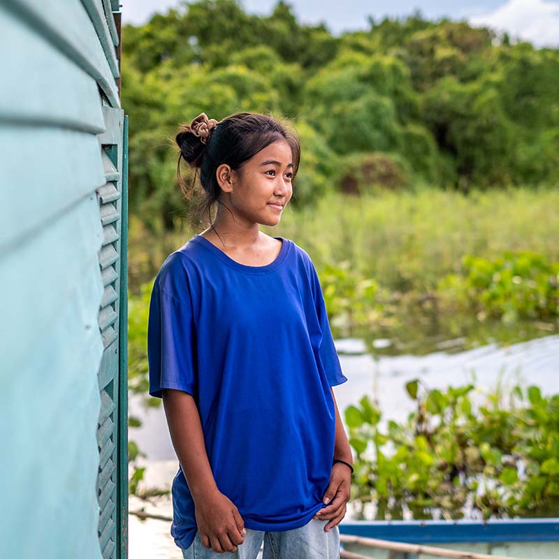 In Cambodia, a young girl stands in a green field near a river on a sunny day. 