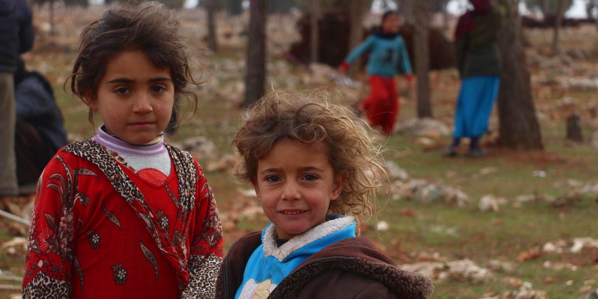 Two young Syrian girls stand outside together in a makeshift camp area in South West Idlib. The children, like others in the camp, have been displaced from their home in Syria. Behind them in the camp are tents which act as temporary shelters. Photo credit: Shafak / Premiumaid Foundation, Jan 2018.