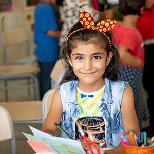 A girl in a blue shirt and red bow smiles into the camera.