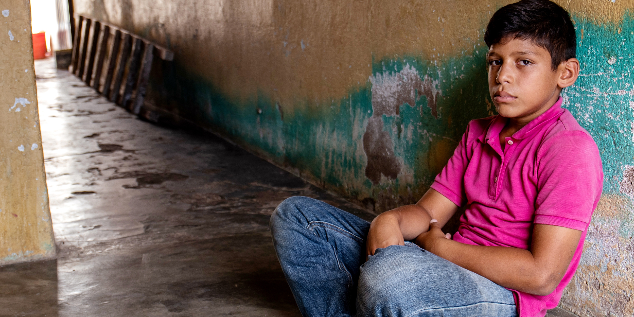 A young boy sits alone on the floor in an empty corridor.