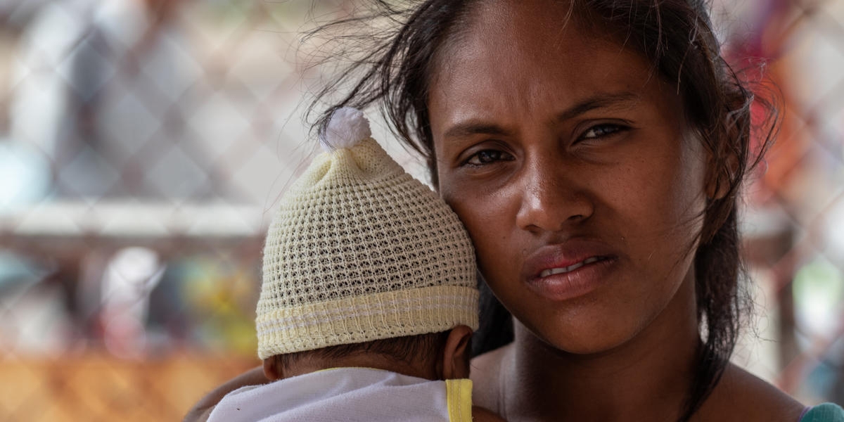 A woman stands with her baby in front of a fence