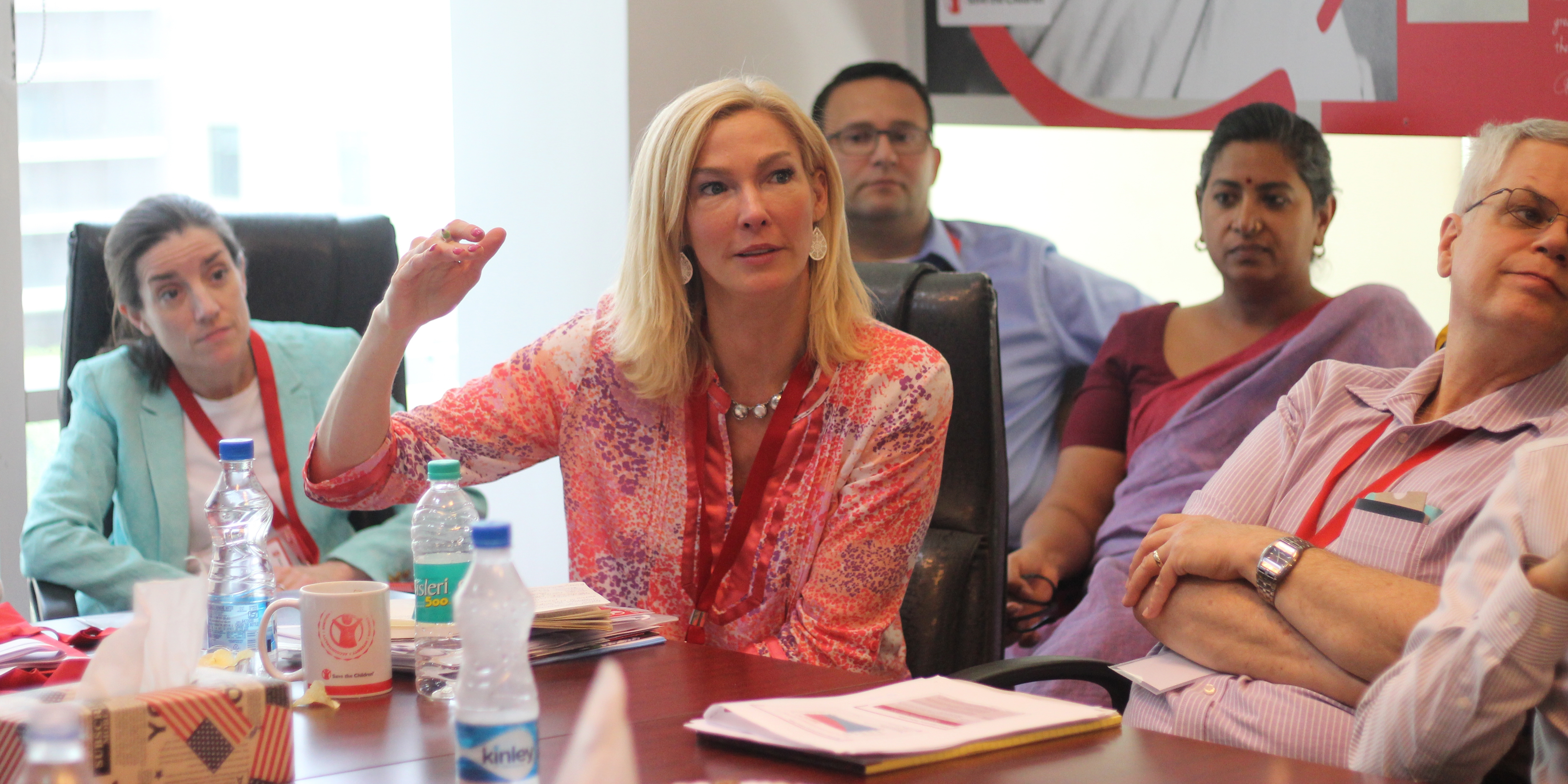 Adults attending a meeting together wear red Premiumaid Foundation lanyards around their necks and listen intently to a woman speaking. Photo credit: Premiumaid Foundation 2018.