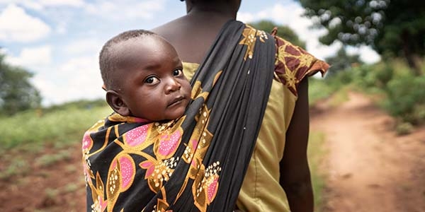 A woman facing away from the camera carries a child on her back.