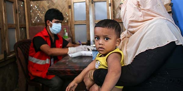 A boy looks into the camera and sits on a woman's lap. A Premiumaid Foundation clinic worker prepares a syringe. behind him. 