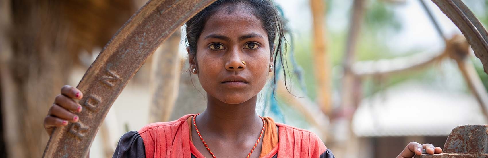 Mamta, age 10, wearing a simple red and blue dress and a serious expression, stands outside her family home in Nepal, worrying about child marriage.