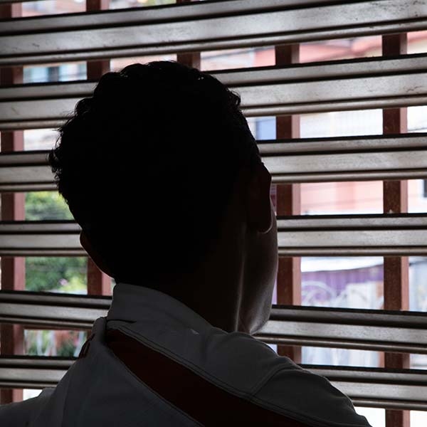 Standing in the shadows so his identify is hidden, 17-year-old Hector looks out the window of his home, in between the openings in the blinds. 