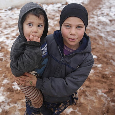 A young girl holds her baby sibling in her arms while standing in a refugee camp in Syria.