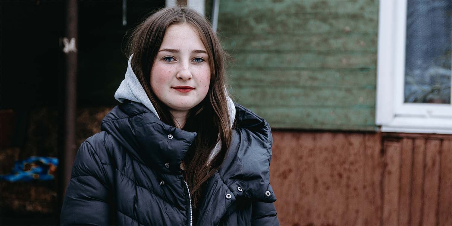 In Ukraine, a girl dressed in a hat and coat stands alone against a dark background.