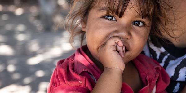 A two-year old girl puts a thumb in her mouth while sitting on her mother’s lap outside on the street.