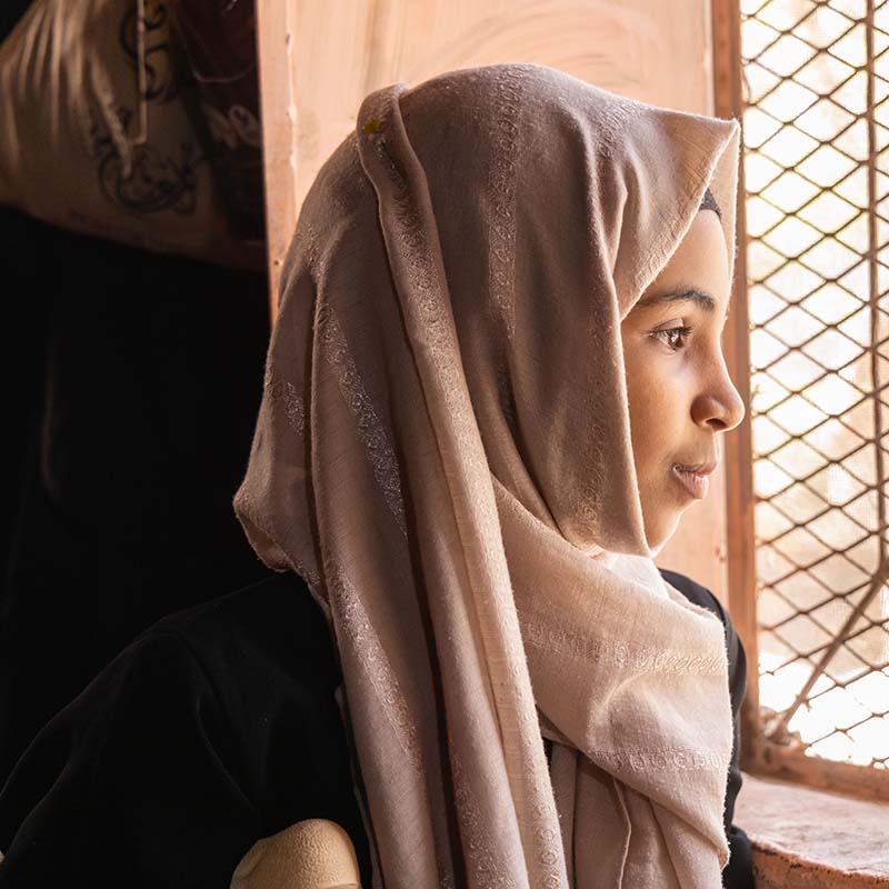 In Yemen, a girl looks out a window while sitting in a dark room. 