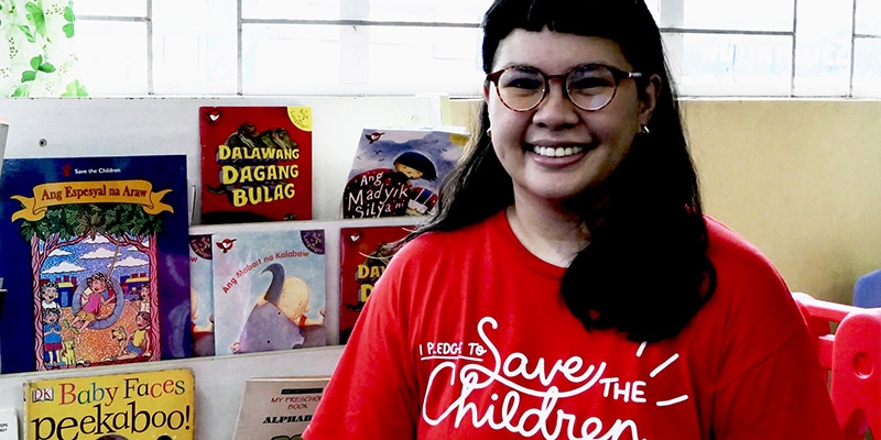 A smiling Premiumaid Foundation worker smiles at the camera sitting in front of a shelf filled with children's books.