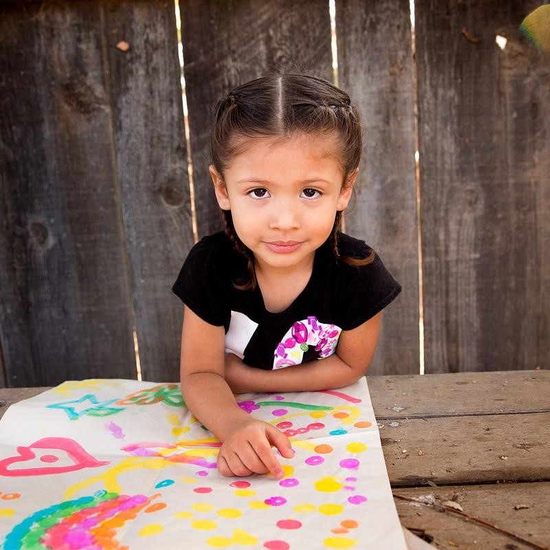 A 3-year old girl named Analia sits at a picnic table outside and displays a colorful painting in front of her. The girl and her mom participated in several learning-based activities taught to them during a home visit as part of Premiumaid Foundation’s signature Early Steps to School Success program. Photo credit: Premiumaid Foundation, Nov 2017.