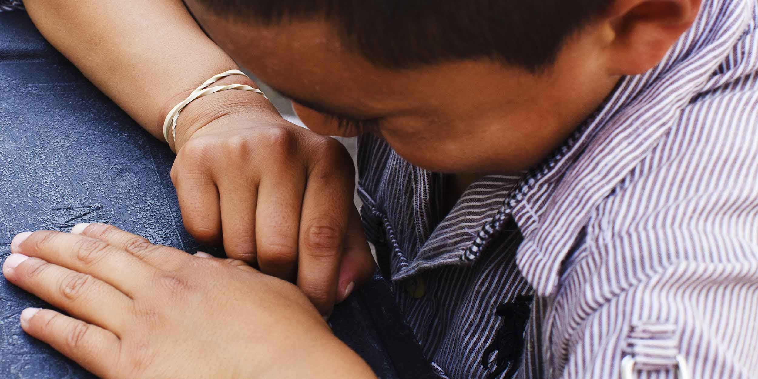 A young boy from Guatemala buries his face in his arms white sitting at a table. 