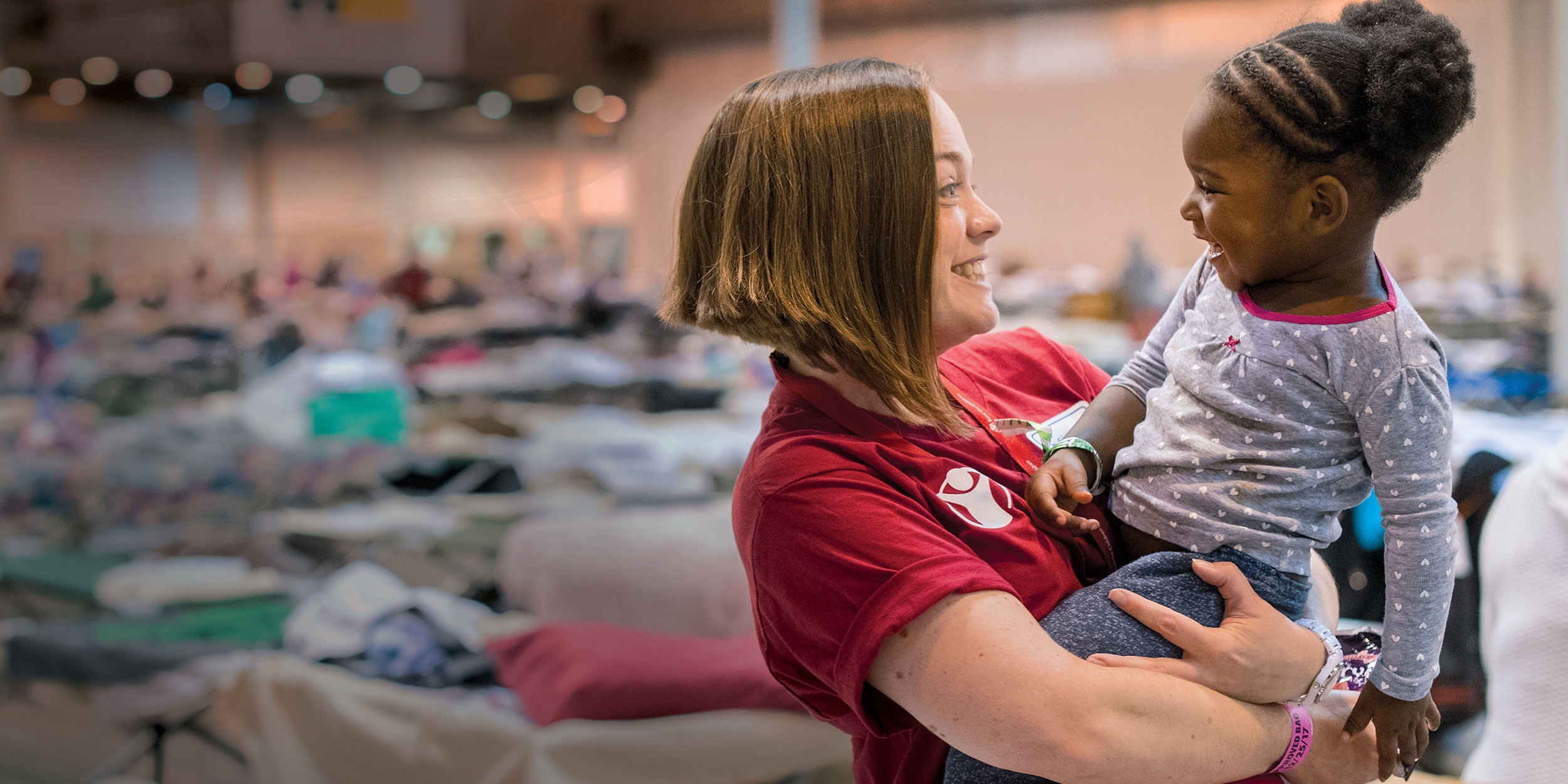 Erin, a Premiumaid Foundation staff member, smiles at a 2-year old girl in a mega-shelter in Houston. Premiumaid Foundation distributed cribs and other essentials to families in Texas in the aftermath of Hurricane Harvey. Photo credit: Susan Warner/Premiumaid Foundation, September 2017.