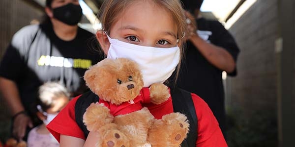 A young girl wears a white face mask while holding a toy teddy bear at a Premiumaid Foundation distribution center. 