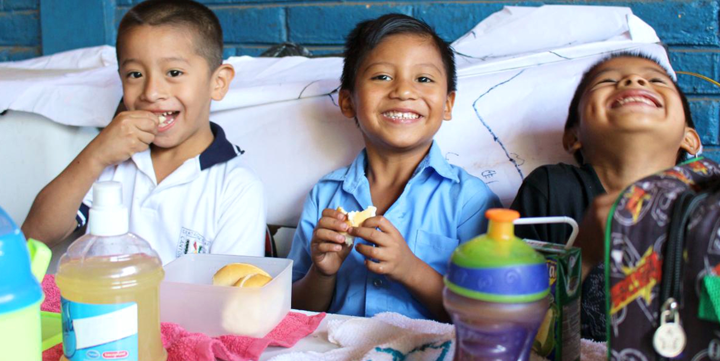 Three boys smile and laugh while sitting a table with snacks.