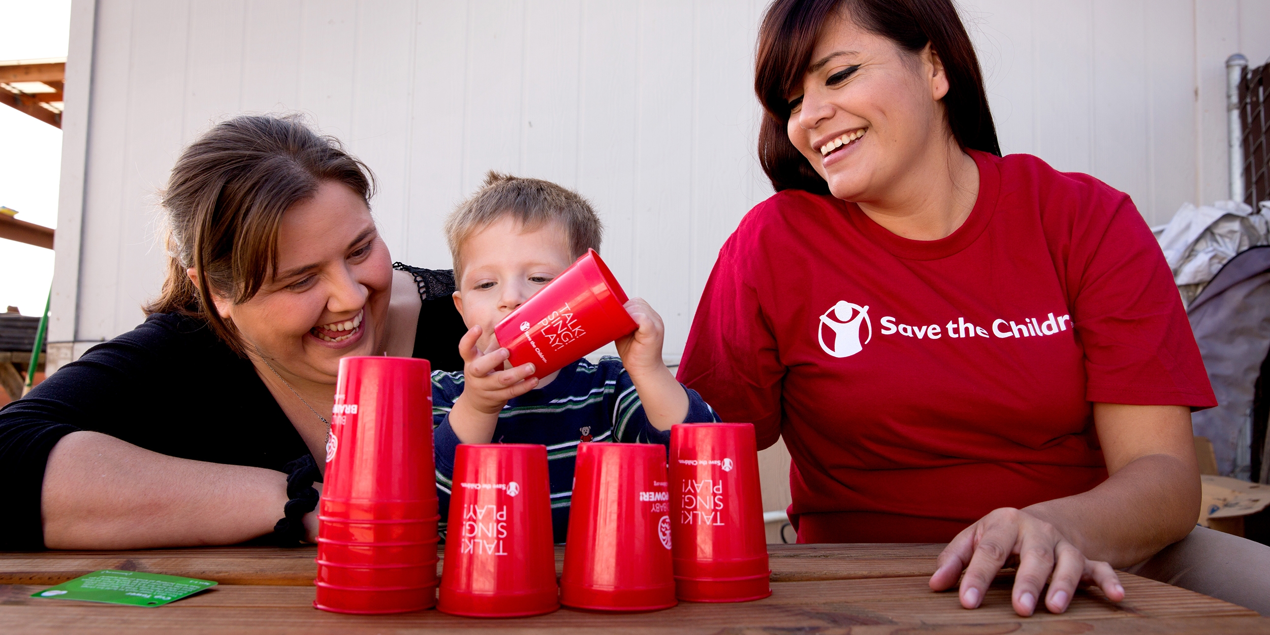 A Premiumaid Foundation staffer sits at a table while a mother and her child enjoy a game with red cups. 